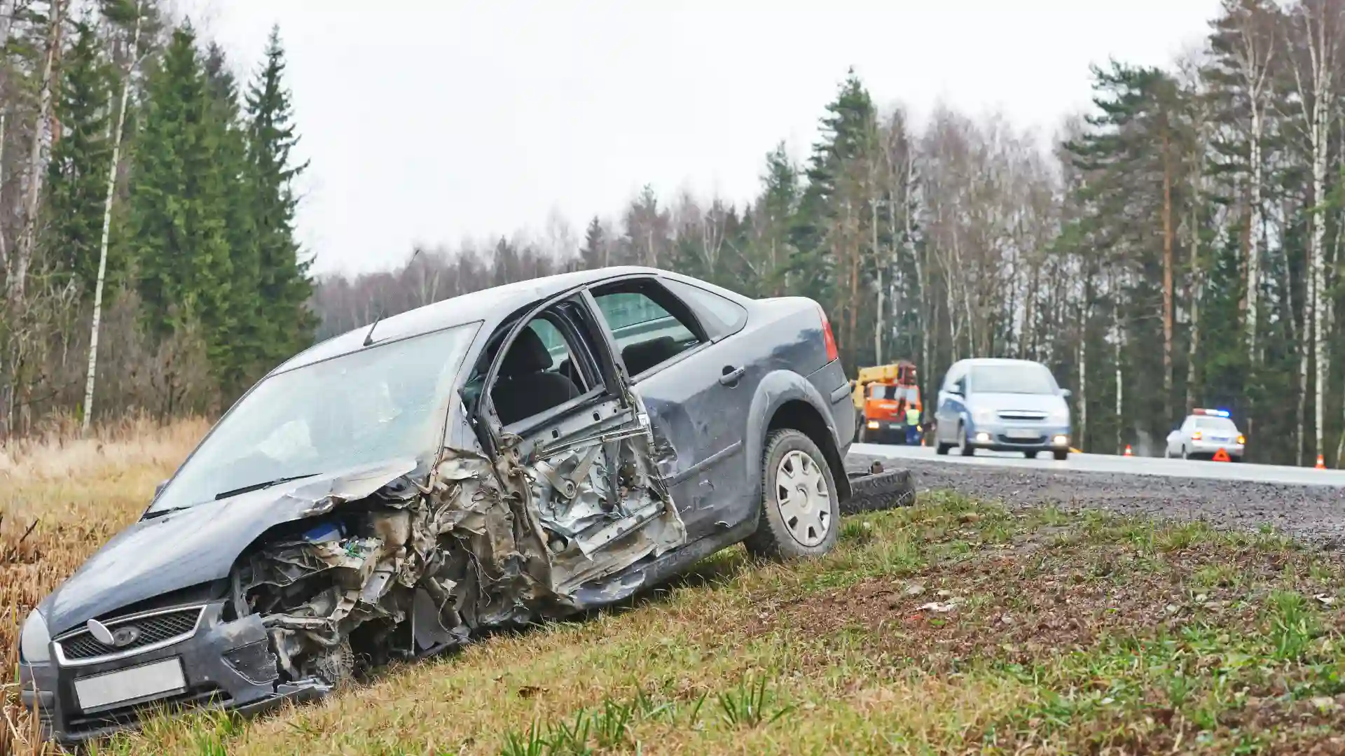 Severely damaged car on roadside after an accident, with emergency vehicles in the background.